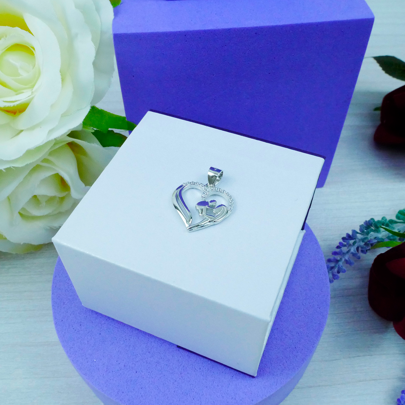 Heart-shaped silver pendant in a white box with purple interior, surrounded by flowers.