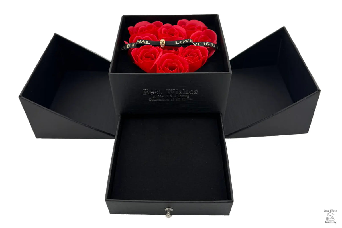 A red jewellery box with a heart-shaped arrangement of red roses on the lid, secured with a magnetic bow. The box is open, revealing cutouts for holding jewelry items like chains, earrings, and rings.