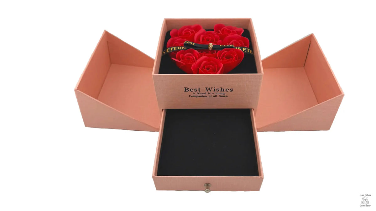 A red jewellery box with a heart-shaped arrangement of red roses on the lid, secured with a magnetic bow. The box is open, revealing cutouts for holding jewelry items like chains, earrings, and rings.