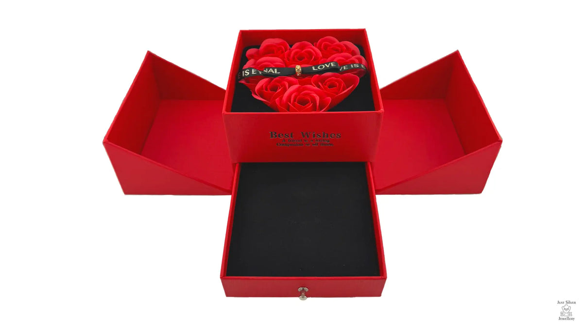 A red jewellery box with a heart-shaped arrangement of red roses on the lid, secured with a magnetic bow. The box is open, revealing cutouts for holding jewelry items like chains, earrings, and rings.