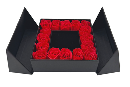 A luxury gift box with a flip-top lid, containing a ring of red roses around a jewelry box with a bow on top.