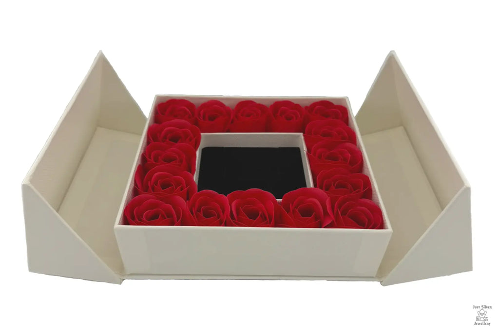 A luxury gift box with a flip-top lid, containing a ring of red roses around a jewelry box with a bow on top.